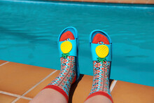 Feet With Colorful Socks And Blue Pool Sandals At The Edge Of A Swimming Pool. Summer Concept. Aesthetic Summer