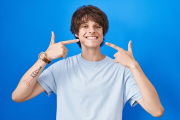 Sticker - Hispanic young man standing over blue background smiling cheerful showing and pointing with fingers teeth and mouth. dental health concept.