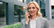 © serg - Portrait of confident and smiling adult caucasian woman in formal clothes walking on street talking on mobile phone