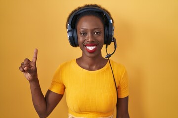 Sticker - African woman with curly hair standing over yellow background wearing headphones showing and pointing up with finger number one while smiling confident and happy.