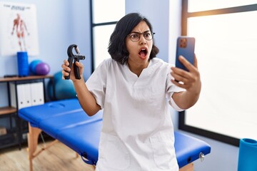 Canvas Print - Young hispanic physiotherapist woman holding hand grip to train muscle doing video call angry and mad screaming frustrated and furious, shouting with anger. rage and aggressive concept.