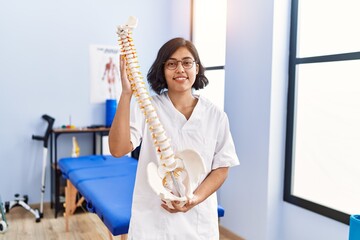 Poster - Young latin woman wearing physiotherapist uniform holding anatomical model of vertebral column at physiotherapy clinic