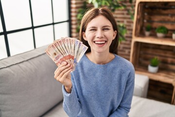 Poster - Young brunette woman holding colombian pesos looking positive and happy standing and smiling with a confident smile showing teeth