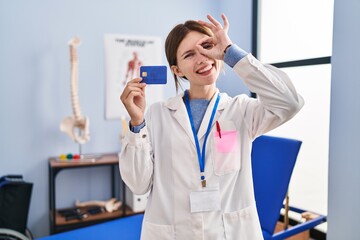 Canvas Print - Young brunette woman working at pain recovery clinic holding credit card smiling happy doing ok sign with hand on eye looking through fingers