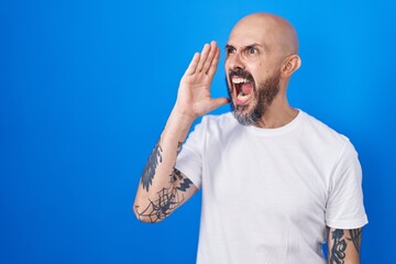 Canvas Print - Hispanic man with tattoos standing over blue background shouting and screaming loud to side with hand on mouth. communication concept.