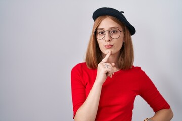 Wall Mural - Young redhead woman standing wearing glasses and beret thinking concentrated about doubt with finger on chin and looking up wondering