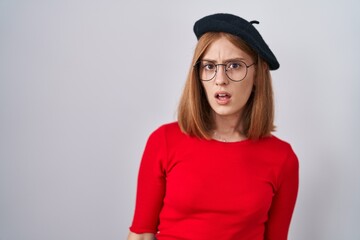 Poster - Young redhead woman standing wearing glasses and beret in shock face, looking skeptical and sarcastic, surprised with open mouth