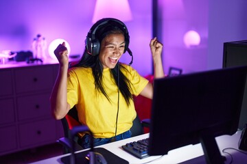 Young beautiful latin woman streamer playing video game with winner expression at gaming room