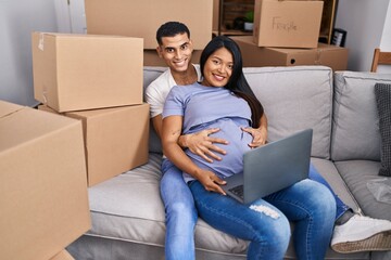 Wall Mural - Young latin couple expecting baby touching belly using laptop at new home