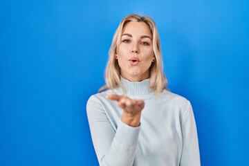 Sticker - Young caucasian woman standing over blue background looking at the camera blowing a kiss with hand on air being lovely and sexy. love expression.