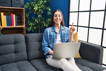 Sticker - Hispanic woman using laptop at home surprised with an idea or question pointing finger with happy face, number one