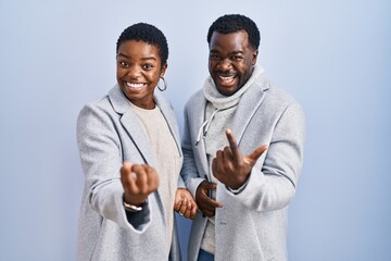 Poster - Young african american couple standing over blue background together beckoning come here gesture with hand inviting welcoming happy and smiling