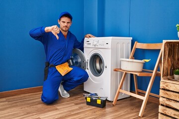 Poster - Young hispanic man working on washing machine with angry face, negative sign showing dislike with thumbs down, rejection concept