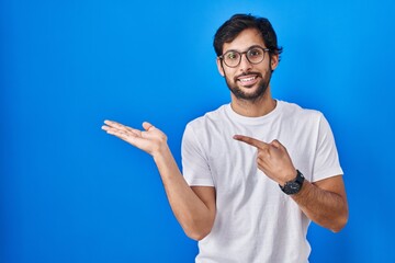 Sticker - Handsome latin man standing over blue background amazed and smiling to the camera while presenting with hand and pointing with finger.