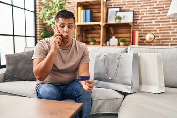 Canvas Print - Hispanic young man doing payment with credit card on the phone making fish face with mouth and squinting eyes, crazy and comical.