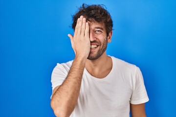 Canvas Print - Hispanic young man standing over blue background covering one eye with hand, confident smile on face and surprise emotion.