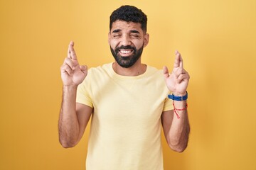 Canvas Print - Hispanic man with beard standing over yellow background gesturing finger crossed smiling with hope and eyes closed. luck and superstitious concept.