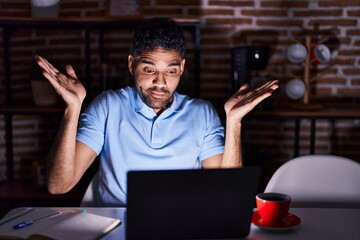 Wall Mural - Hispanic man with beard using laptop at night clueless and confused expression with arms and hands raised. doubt concept.