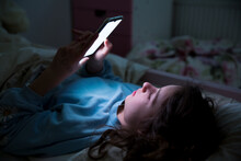 A Child Using Smart Phone Lying In Bed Late At Night, Playing Games, Watching Videos Online, Scrolling Screen. Children's Screen Addiction And Parent Control Concept. Child's Room At Night. 