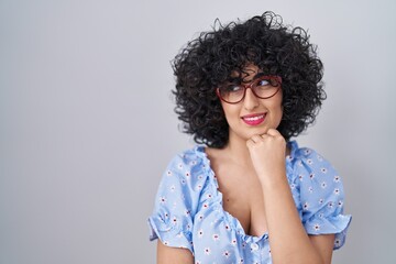 Poster - Young brunette woman with curly hair wearing glasses over isolated background looking confident at the camera smiling with crossed arms and hand raised on chin. thinking positive.