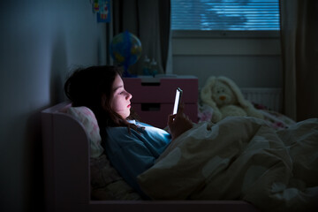 A child using smart phone lying in bed late at night, playing games, watching videos online, scrolling screen. Children's screen addiction. Child's room at night. 