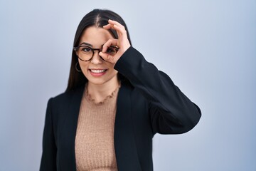 Poster - Young brunette woman standing over blue background doing ok gesture with hand smiling, eye looking through fingers with happy face.