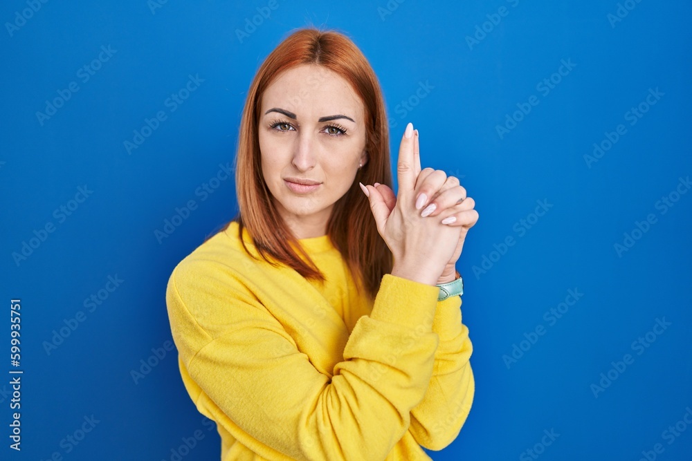 Young woman standing over blue background holding symbolic gun with hand gesture, playing killing shooting weapons, angry face