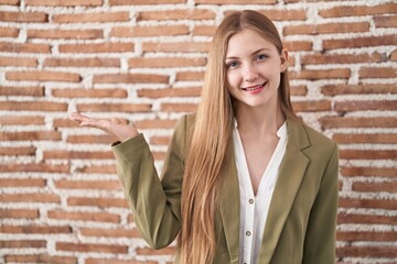 Canvas Print - Young caucasian woman standing over bricks wall background smiling cheerful presenting and pointing with palm of hand looking at the camera.