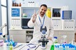 © Krakenimages.com - Young hispanic man with beard working at scientist laboratory smiling looking to the camera showing fingers doing victory sign. number two.