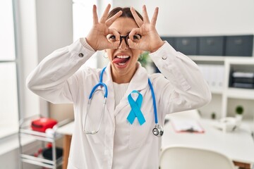 Canvas Print - Young brunette doctor woman wearing stethoscope at the clinic doing ok gesture like binoculars sticking tongue out, eyes looking through fingers. crazy expression.