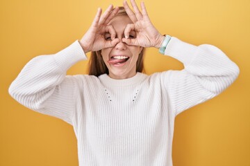 Sticker - Young caucasian woman wearing white sweater over yellow background doing ok gesture like binoculars sticking tongue out, eyes looking through fingers. crazy expression.