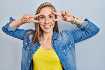 Poster - Young blonde woman standing over blue background doing peace symbol with fingers over face, smiling cheerful showing victory