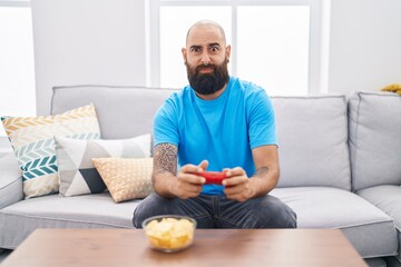 Canvas Print - Young hispanic man with beard and tattoos playing video game sitting on the sofa clueless and confused expression. doubt concept.