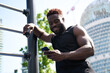 © insta_photos - Happy fit sporty young black man standing near bar in workout park holding phone using mobile trainings app. Strong African ethnic guy bodybuilder looking at smartphone in outdoor gym.
