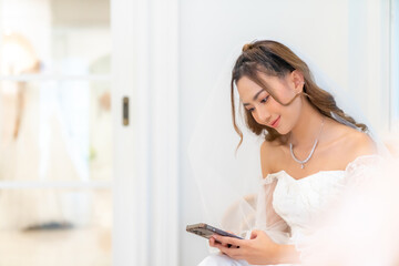 Portrait of Beautiful Asian woman bride in white wedding dress using mobile phone in fitting room at bridal shop. Wedding ceremony marriage concept and wireless technology concept.