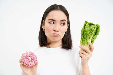 Healthy lifestyle concept. Young woman deciding between healthy vegetables, cabbage and delicious glazed doughnuts, white background