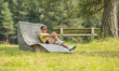 © Dirk - Caucasian man sitting on a design wooden bench in a meadow in Austria. Pine trees are standing in the background. Summer, day, resting, relaxing.