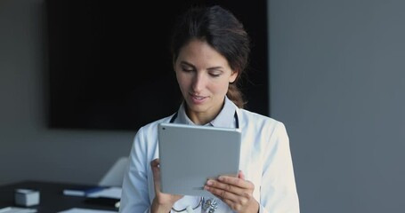 Wall Mural - Smiling attractive Latina female doctor in white medical uniform use tablet gadget in modern private hospital, looking at pad screen, consult client on-line, fill patient medical history or anamnesis