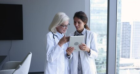 Wall Mural - Young and mature female physicians talking standing in clinic office, discuss patient treatment plan, disease and symptoms using modern device tablet. Teamwork of intern and medical chief in hospital