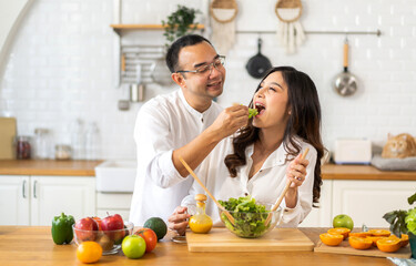 Young asian family couple having fun cooking and preparing cook vegan food healthy eat with fresh vegetable salad on counter in kitchen at home.Happy couple looking to preparing food