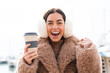 Young woman wearing winter muffs  and holding take away coffee at outdoors with shocked facial expression