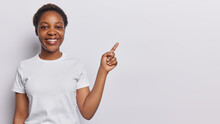 Studio Shot Of Pretty Dark Skinned Millennial Girl With Short Hairstyle Points Index Finger Above On Blank Space Or At Advertisement Area Discusses Amazing Promo Dressed In Casua White L T Shirt