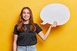 © WHstudio Leushin N - Waist up shot of satisfied millennial girl holds blank speech bubble smiles broadly suggests to place your advertising content here dressed in black t shirt and jeans isolated over yellow background