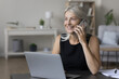© fizkes - Cheerful happy middle aged entrepreneur woman talking on cellphone at table with laptop, using wireless online connection technology for work from home, smiling, enjoying mobile phone conversation