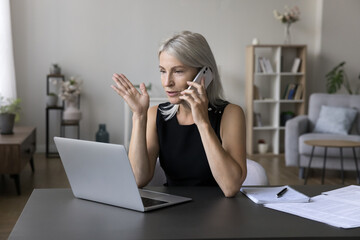 positive busy mature freelance business woman speaking on mobile phone at home workplace, sitting at