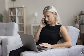 Focused senior freelance professional woman speaking on cell, talking on telephone call, using online app on laptop for distant job, making order, sitting in home armchair