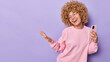 © wayhome.studio  - Joyful curly haired woman feels boost of happiness foolishes around holds mobile phone looks gladfully aside smiles broadly wears spectacles and casual pullover isolated over purple background