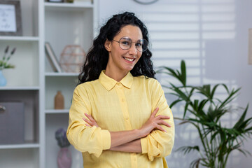 Portrait of young beautiful hispanic at home, woman with curly hair in glasses and yellow shirt smiling and looking at camera, successful businesswoman working inside home office with crossed arms.