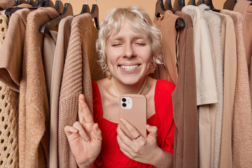 Beautiful happy blonde woman keeps fingers crossed believe in good luck searches clothes on clothing rack holds mobile phone wears red dress and necklace. People and shopping online concept.