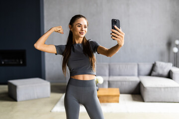 Portrait of sportive powerful woman dressed in tight gray top demonstrating her biceps taking selfie or having video call at home